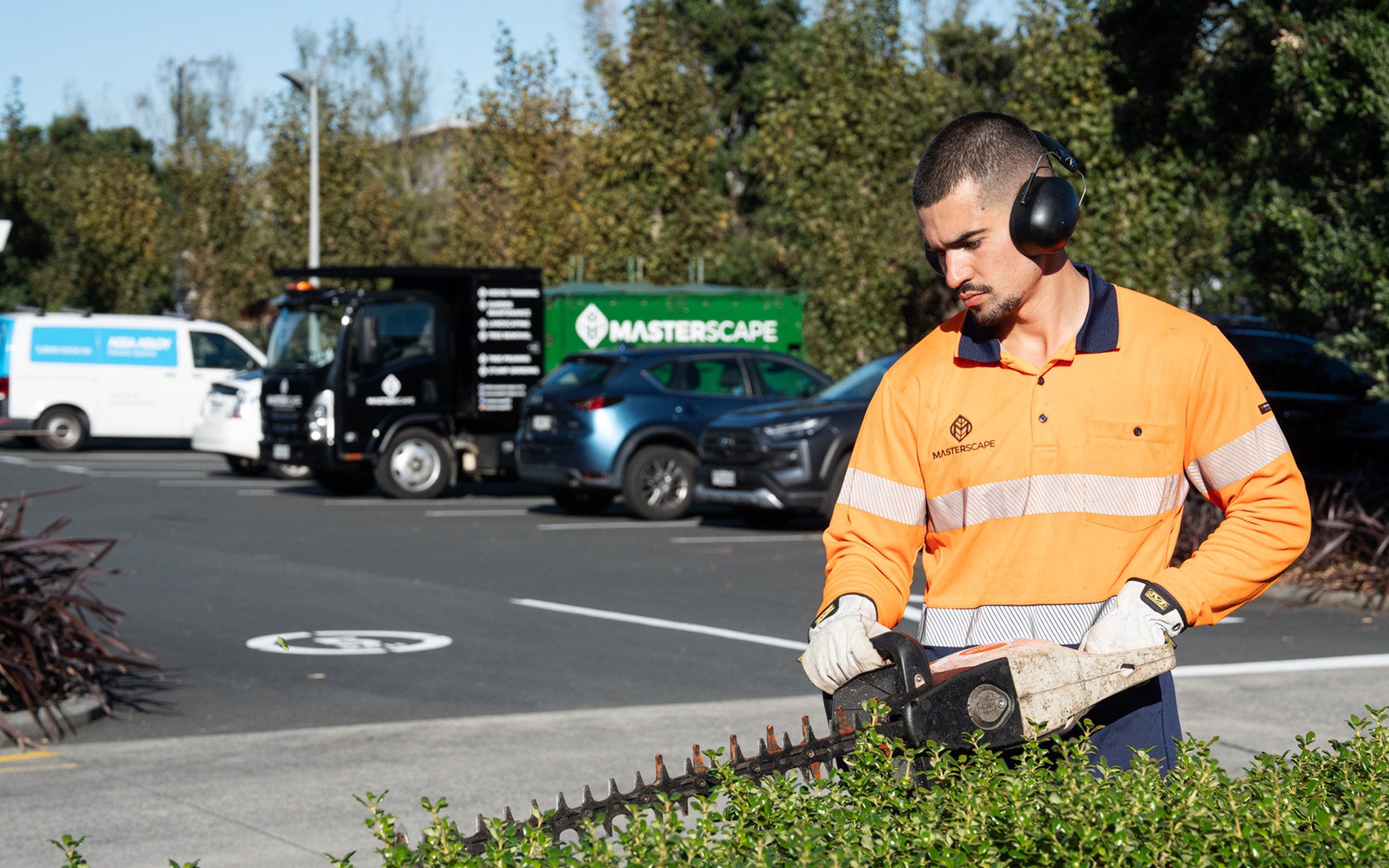 Masterscape crew member trimming hedges in a commercial car park with branded Masterscape truck visible in the background in Auckland